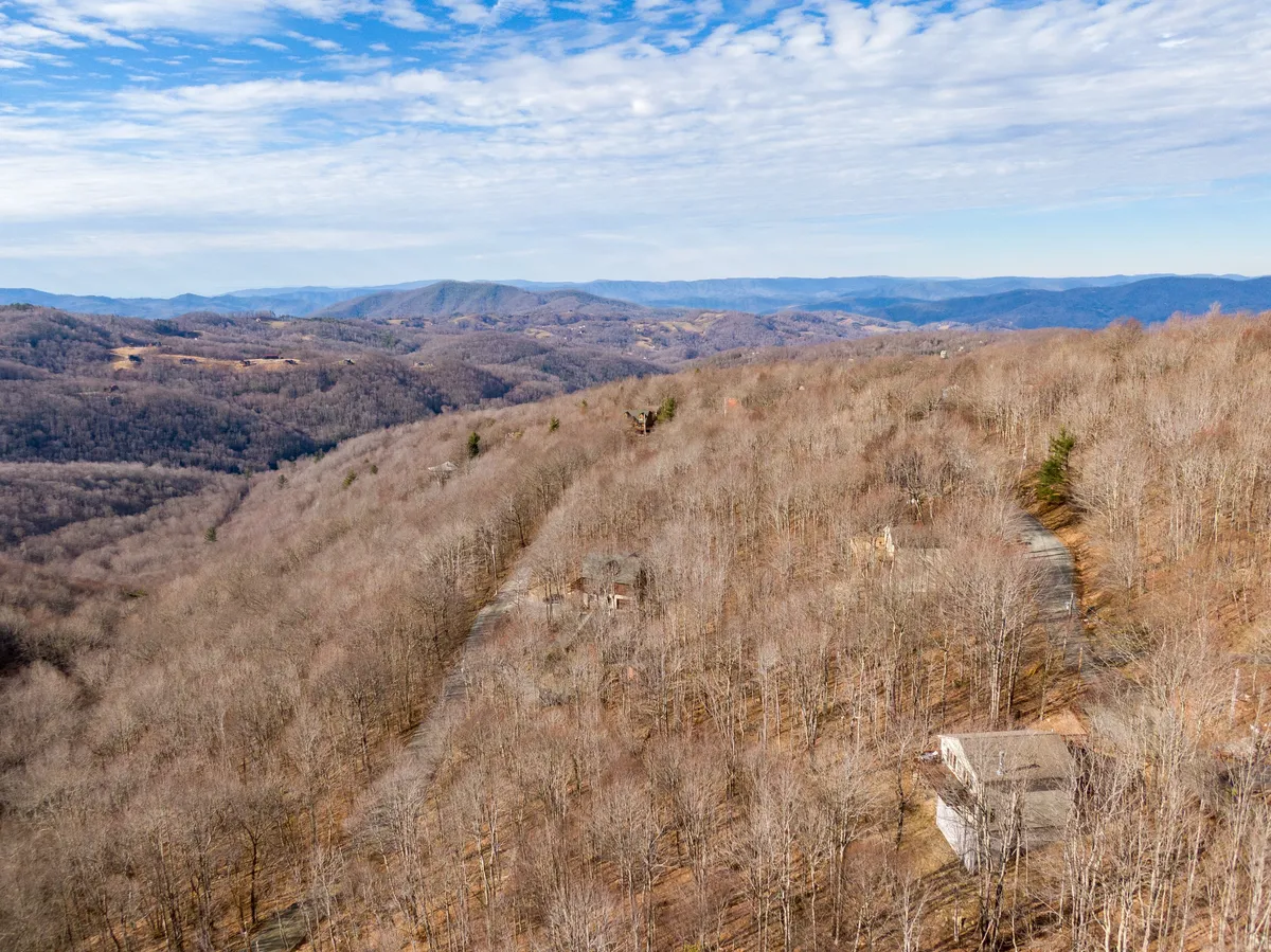 The image showcases a breathtaking, unobstructed vista of rolling hills and distant Blue Ridge mountains under a vast sky, with bare winter trees framing the view and occasional mountain homes dotting the landscape. The serene scene suggests quiet mornings and tranquil evenings, with sunrise light casting a warm glow across the slopes.