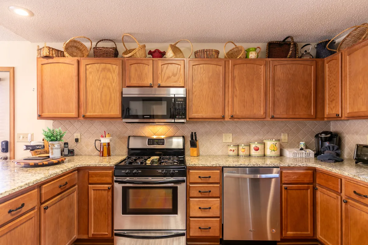 A bright, open kitchen featuring stainless steel appliances, granite countertops, warm wooden cabinetry, and decorative baskets. Ample counterspace and modern amenities are visible, conveying a sense of readiness for both everyday cooking and special gatherings.