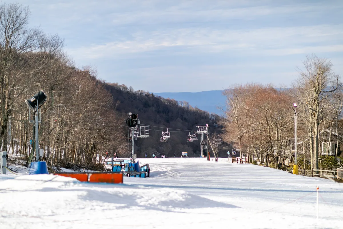 A wide shot of a ski lift ascending a snow-covered slope, framed by tall trees and an open sky. The scene evokes excitement and the allure of mountain exploration, suitable for all ages and skill levels.