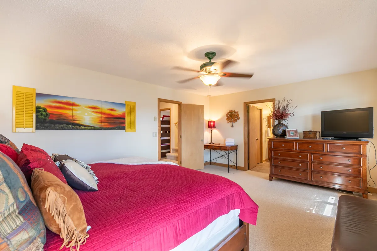 A cozy bedroom with a bed dressed in a red quilt, a sunset painting above the wooden dresser, a television, and cheerful yellow shutters. The room feels warm and offers entertainment options for winding down.