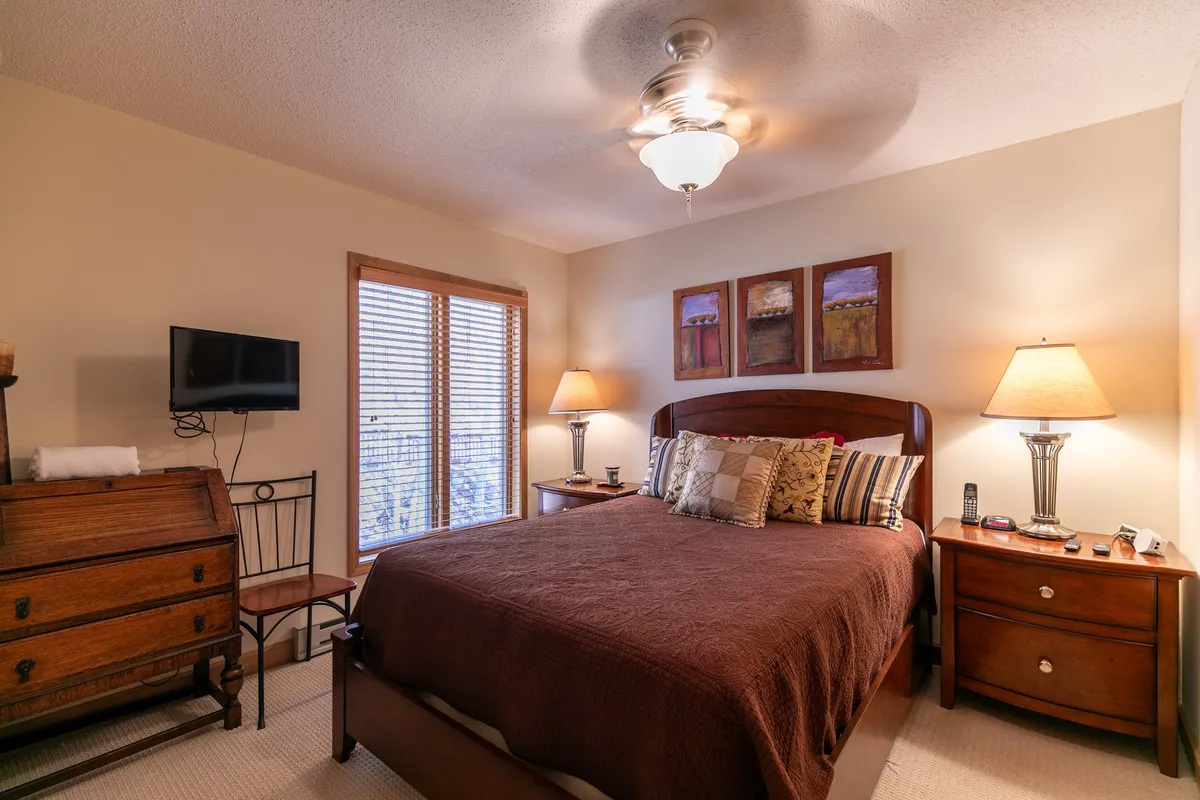 A warmly lit bedroom featuring a plush bed with a brown quilt, two bedside lamps, paintings on the wall, and a wooden dresser. The overall atmosphere is serene and inviting, ideal for relaxation and privacy.