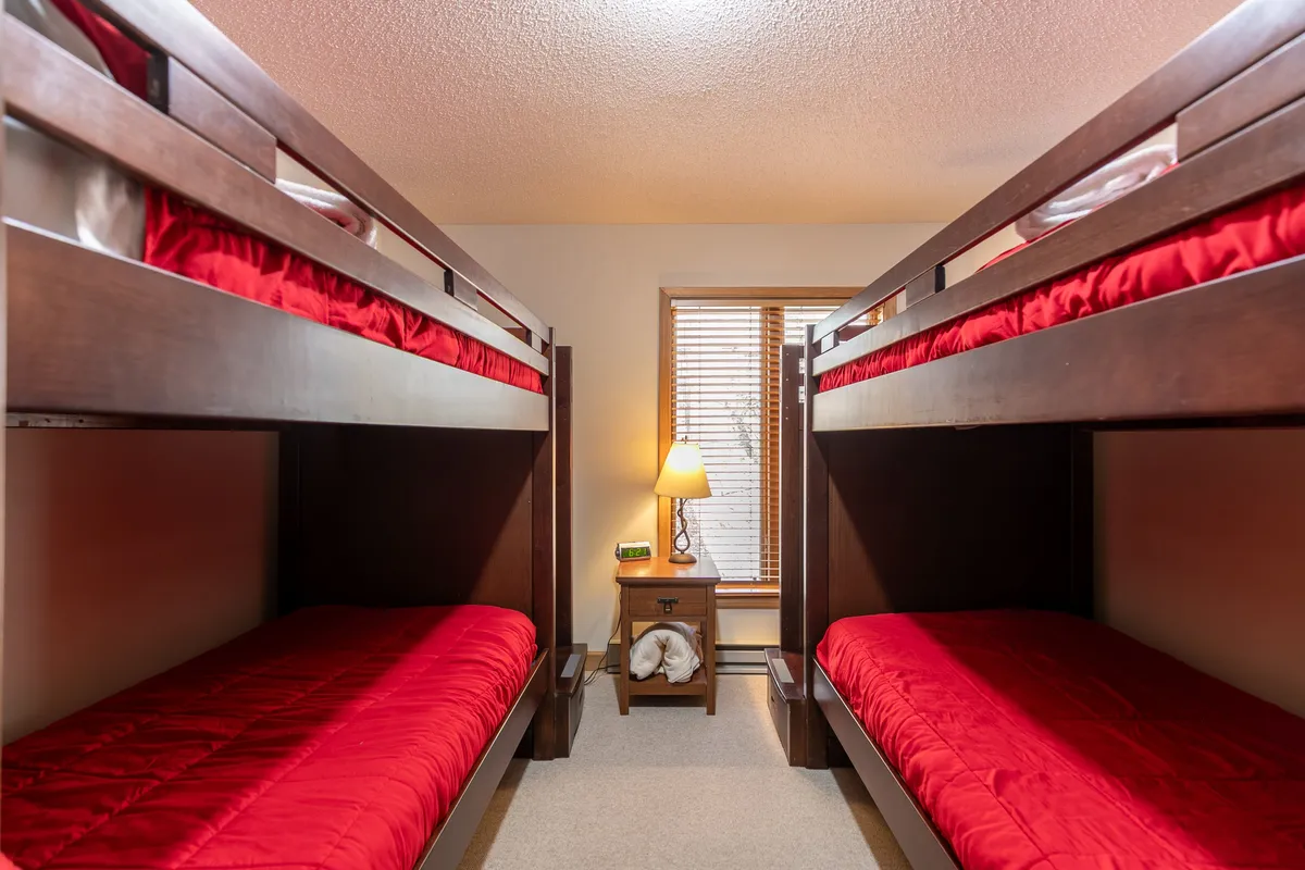 A playful and bright bunk room featuring two sets of twin-over-twin beds, a nightstand, and a window with blinds. The arrangement encourages camaraderie and fun for kids, with plenty of space for sleep and imaginative play.