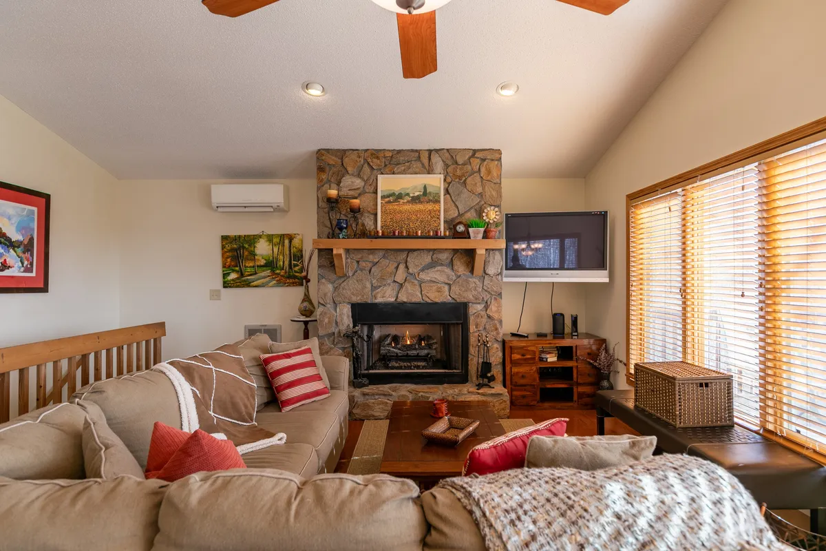 A warm, welcoming living room centered around a stone fireplace, comfortable sofa with colorful cushions, and large windows that flood the space with natural light. The décor evokes a cozy, homey atmosphere, perfect for unwinding or hosting friends and family. The view through the windows reinforces a connection to the outdoors.