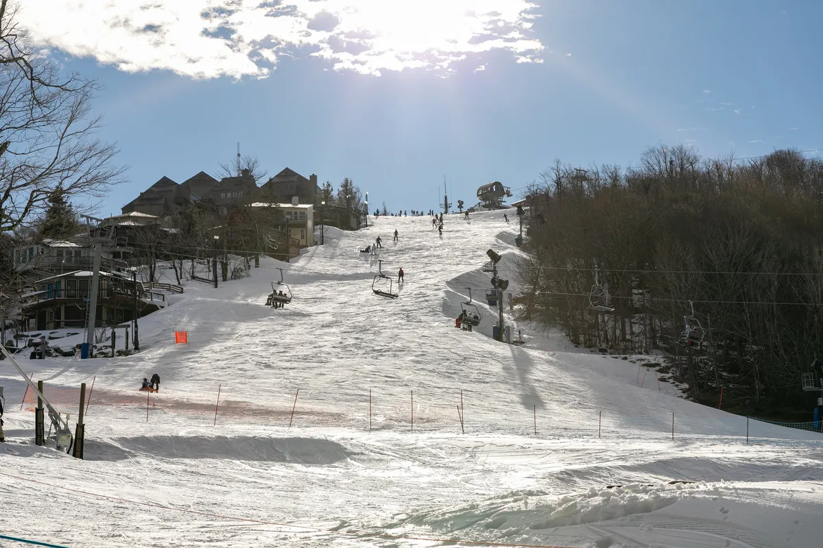 A lively exterior winter scene featuring ski slopes bustling with people skiing and snowboarding, a ski lift in operation, and surrounding mountain cabins nestled among snow-covered trees. The sky exhibits late afternoon light, enhancing the excitement and adventure of winter activities.