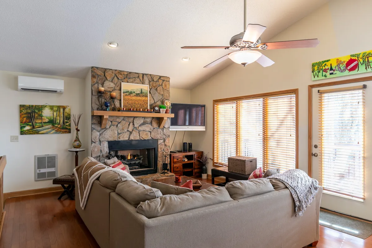 A cozy living room featuring a prominent stone fireplace, large windows that frame scenic outdoor views, and a comfortable sofa. The room feels inviting and perfect for relaxation or intimate conversations, with natural light enhancing its warmth.
