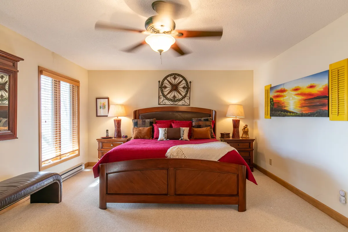 A restful bedroom with red bedding, tasteful wall art, and wood furnishings. Large windows with blinds invite in natural light and provide the opportunity to wake up to scenic views, enhancing the sense of tranquility and privacy.