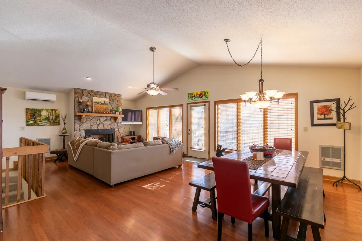 A wide view of the open-concept living space featuring a stone fireplace, a spacious dining table, large windows framing mountain vistas, and a cozy sofa. The room is filled with warm, natural light and set up for gatherings or quiet evenings by the fire.